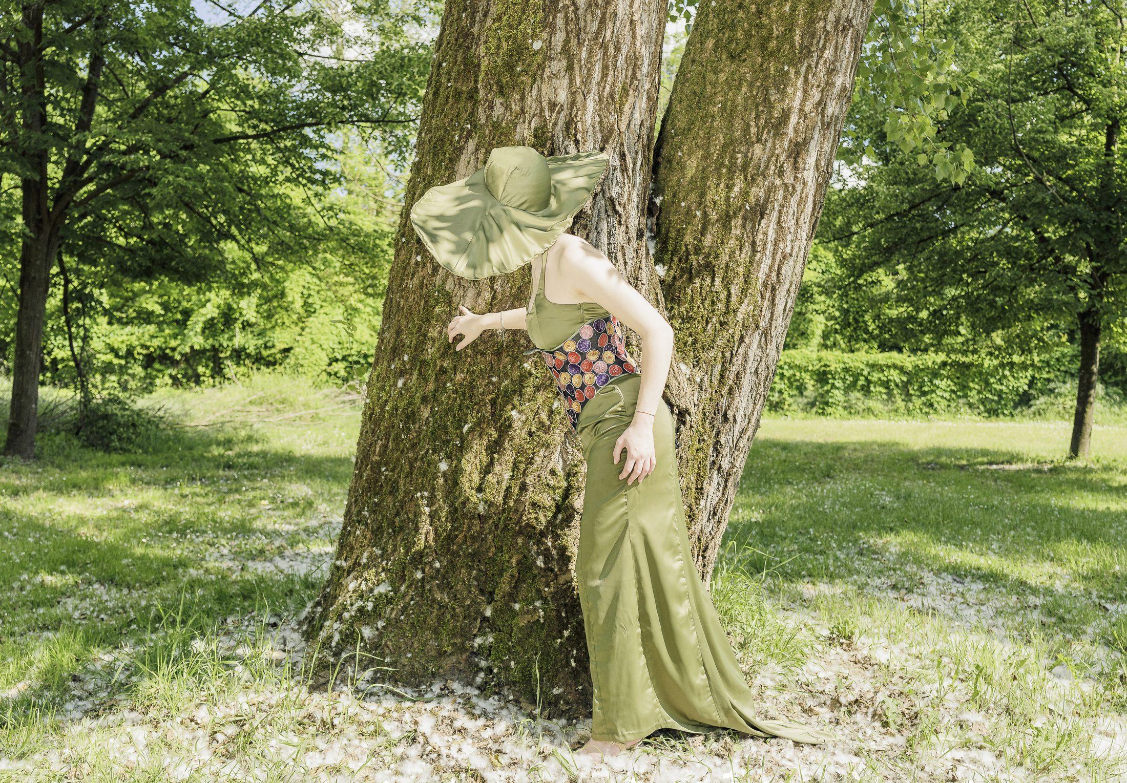 Abito lungo verde oliva con cappello a tesa larga, modella chinata contro il tronco di un grande albero