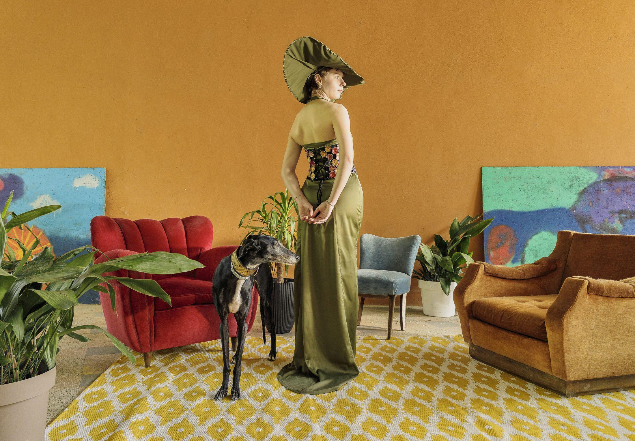 Abito lungo in raso verde oliva con cappello a tesa larga, in un salotto color ocra con poltrone d’epoca