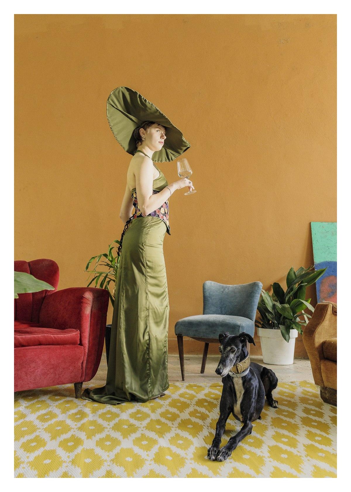 Abito lungo verde oliva e cappello a tesa larga, modella di profilo in un salotto ocra con un levriero