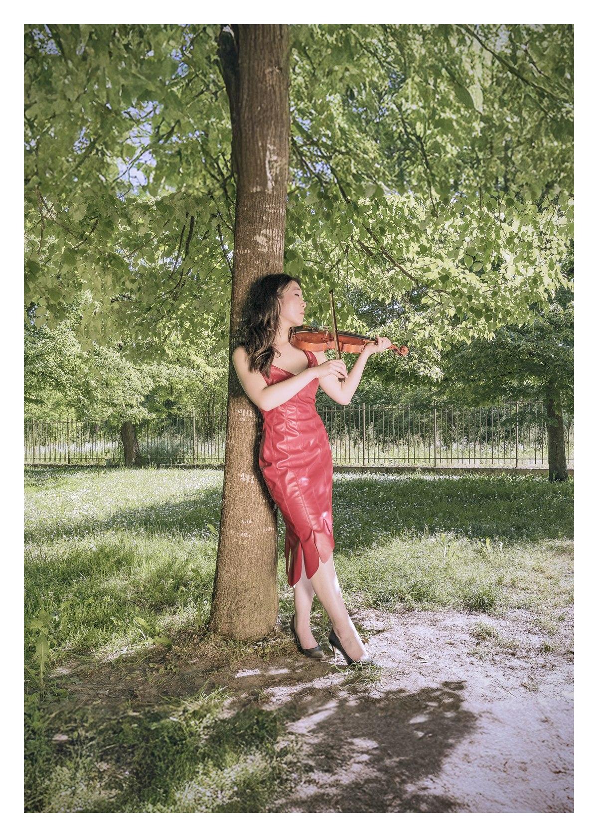 Modella in tubino rosso con orlo a punte mentre suona il violino accanto a un albero nel parco